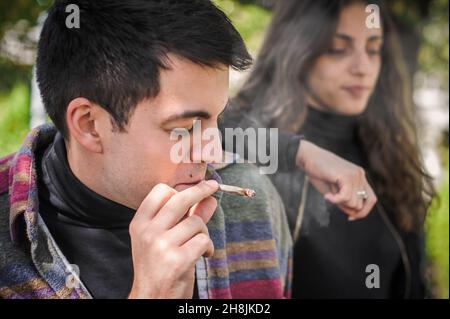 Young relaxed and smiling happy young couple, woman and man, smoking cannabis marijuana ganja or hashish joint in public park outdoor. Female and male Stock Photo