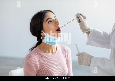 Nurse in gloves with protective equipment performs coronavirus swab from mouth on millennial indian lady in clinic Stock Photo