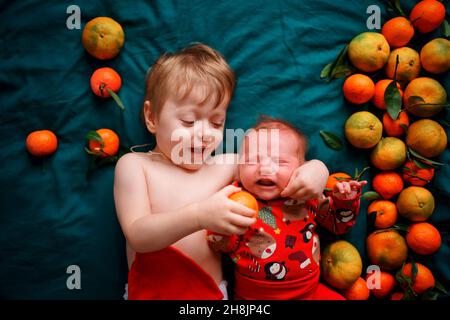 The older brother wants to feed the Christmas newborn baby a tangerine, the baby cries. Children lie on a green blanket next to tangerines. Stock Photo