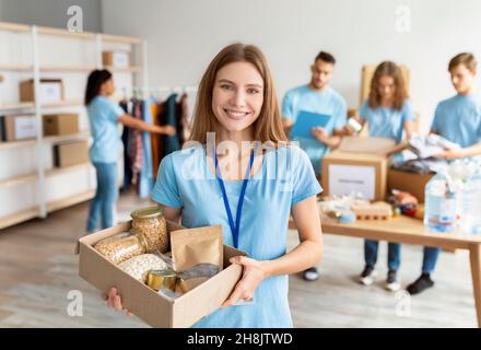 Positive female volunteer in blue uniform holding cardboard box with food donation, working in charity center Stock Photo