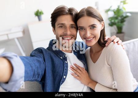 Smiling young european lady and guy hugging, look at webcam and take selfie on living room interior Stock Photo