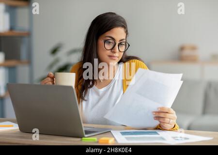 Home office. Concentrated arab businesswoman working with papers and laptop, holding cup with hot beverage Stock Photo