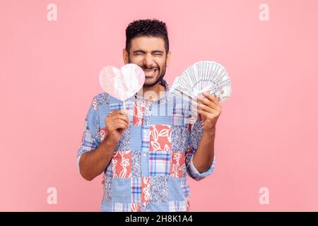 Happy satisfied brunette man in blue casual style shirt holding paper heart and dollar banknotes, standing with closed eyes and toothy smile. Indoor studio shot isolated on pink background. Stock Photo
