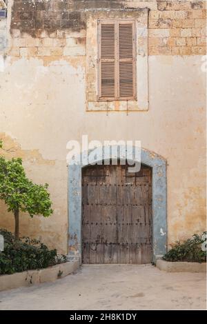 MDINA, MALTA - Jul 24, 2021: Decaying wooden door, large headed nails as ornament, flaking paint on jambs, decaying rectangular, louvred window above. Stock Photo