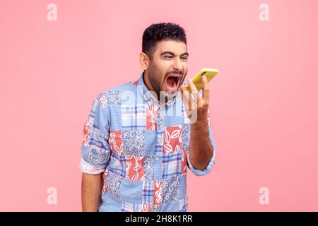 Portrait of angry mad man with beard in blue casual shirt loudly screaming talking phone, leaving voice message with furious aggressive expression. Indoor studio shot isolated on pink background. Stock Photo