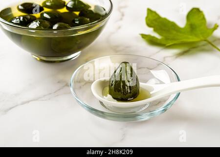 Close-up of preserved whole unripe fig on a spoon near bowl of delicious green figs in syrup jam over marble surface. Traditional mediterranean food. Stock Photo