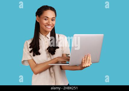 Attractive woman blogger working on new post for her blog on laptop, looking at camera with happy inspired smile, watching funny video on internet. Indoor studio shot isolated on blue background. Stock Photo