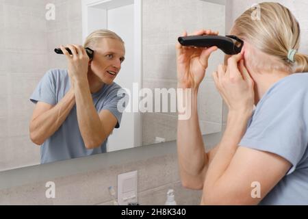 Man shaving whiskers hair by himself, self haircut at home in front of mirror in the bath. Male with long haircut using shaving machine trimmer Stock Photo