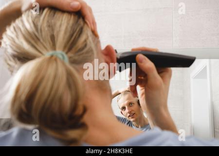 Man shaving whiskers hair by himself, self haircut at home in front of mirror in the bath. Male with long haircut using shaving machine trimmer Stock Photo