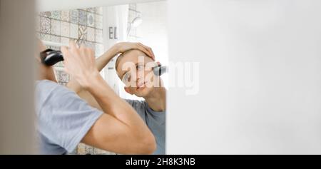 Man shaving whiskers hair by himself, self haircut at home in front of mirror in bath. Male with long haircut using shaving machine trimmer. Electric Stock Photo