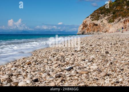 Sunny blue Mylos pebble beach with azure sea, white foam and rocky cliff on coast of Lefkada island in Greece. Summer nature vacation travel to Ionian Stock Photo