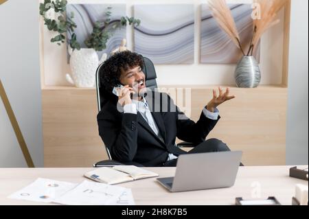 Busy successful Indian businessman, IT specialist, company owner, sitting at work desk at home, having a pleasure cell phone conversation during work time, smile friendly, looks away Stock Photo