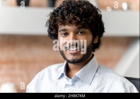 Close-up portrait of Indian young adult positive curly haired male office worker, wearing shirt, looking straight into camera with pleasure smile Stock Photo