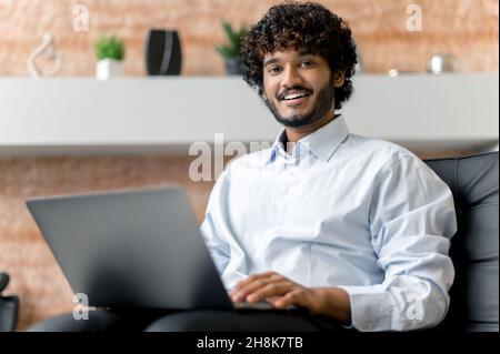Indian positive confident curly-haired young adult guy, freelancer or manager in shirt, sitting on armchair with laptop looking at the camera and smiling friendly Stock Photo