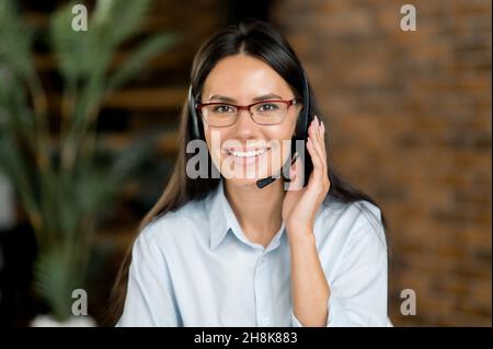 Close-up headshot of a pleasant, confident brunette caucasian woman in headset, freelancer, call center worker or consultant, wearing formal stylish shirt, looks directly at camera, smiles friendly Stock Photo