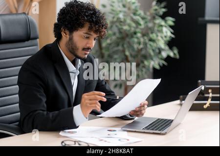 Serious focused successful Indian businessman in formal suit, real estate or stock market agent, sitting at his workplace in office, studying financial charts, analyzing profit, growth dynamics Stock Photo