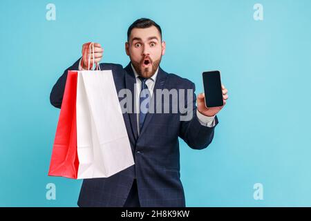 Surprised bearded man wearing official style suit holding in hands smartphone with blank display and shopping bag, shocked with easy shopping online. Indoor studio shot isolated on blue background. Stock Photo