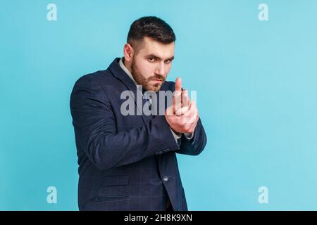 Hey, I'll kill you. Bearded businessman in suit pointing with finger pistols to camera and threatening to shoot, pretending to be dangerous criminal. Indoor studio shot isolated on blue background. Stock Photo