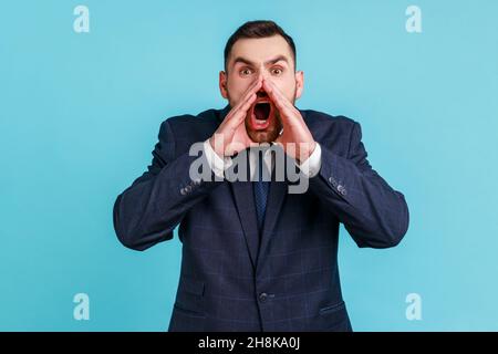 Portrait of hysterical young adult man wearing official style suit loudly yelling in panic, holding hands near his mouth, teenager protest. Indoor studio shot isolated on blue background. Stock Photo