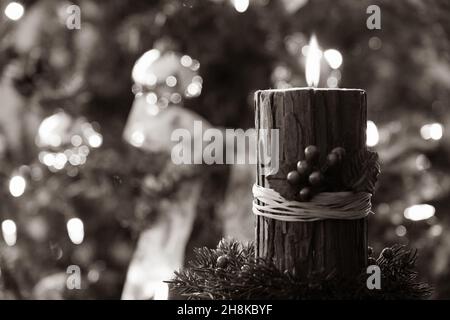 Christmas candle with red berries and rustic bark wrap lit in a dark room in front of a decorated Christmas tree Stock Photo