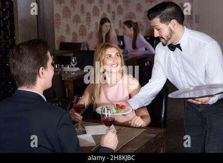 Polite waiter bringing ordered dishes to smiling couple at restaurant Stock Photo