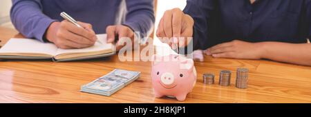 saving concept the man in blue sweater writing income account while the female in dark blue shirt drop a coin into a piggy bank. Stock Photo