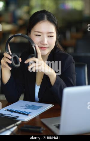 Beautiful asian businesswoman holding headphone while looking at laptop screen at her workplace. Stock Photo