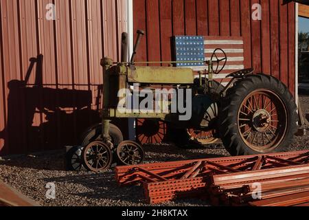 An old rusty green tractor in front of a red barn with a home made wooden American flag on the wall. Stock Photo