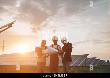 African american technician, muslim woman and indian man standing together on solar farm and looking on blueprints. Multiracial people planning strategy of alternative energy production. Stock Photo