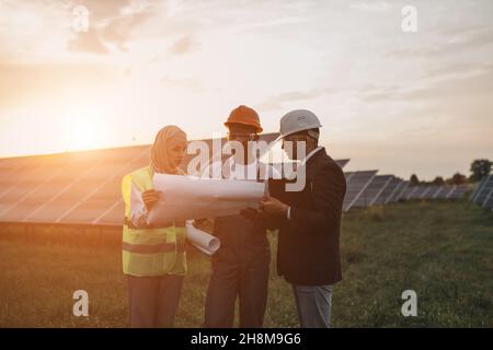 Group of three multiracial people in safety helmets having working meeting among solar station. Technician with engineers standing outdoors with blueprints in hands. Stock Photo