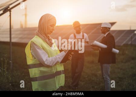 Side view of muslim woman in hijab writing on clipboard while standing among rows of solar panels. Two multicultural men in helmets standing behind with project plan in hands. Stock Photo