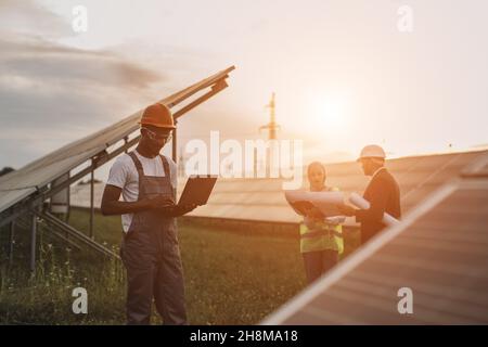 African american technician in uniform standing on field with solar panels and typing on laptop while muslim woman and indian man working with blueprints behind. Alternative energy concept. Stock Photo