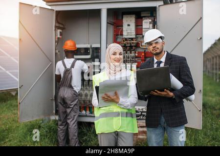 Muslim woman in hijab and indian man in suit using laptop and clipboard during discuss at solar station. African american technician working with cables in transformer on background. Stock Photo