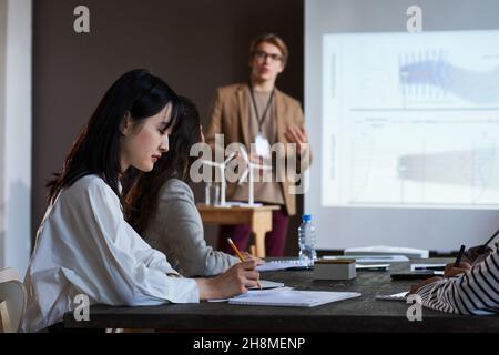 Asian young businesswoman making notes while sitting at the table with her colleagues during business seminar Stock Photo