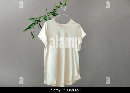 Beige T shirt hanging on hanger on plant Bamboo branches Isolated on gray background. Minimalistic Eco Concept Basic clothing T-shirt mockup. Stock Photo