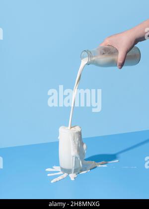 Woman hand pouring milk from a bottle into the glass, minimalist on a blue background. Over-spilling milk on the blue-colored table. Stock Photo