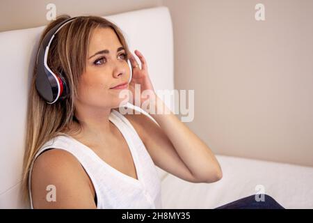Woman listening to music with headphones sitting on the bed in her room. Girl with closed eyes. Enjoy the music. Stock Photo