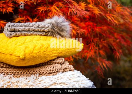 Stack of knitted warm clothing and wool pillow. Autumn leaf background. Knit hat with pom pom and sweater Stock Photo