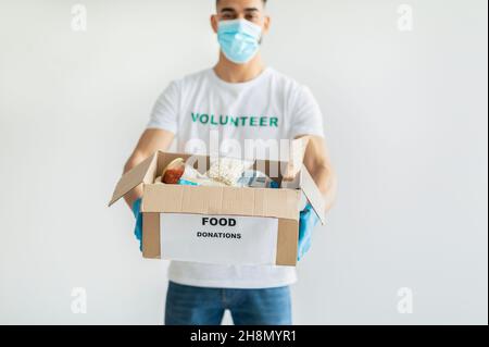 Food donations. Millennial arab man in volunteer t-shirt and medical mask, showing box with canned food and packages Stock Photo