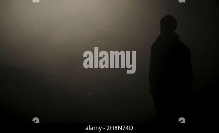 Black silhouette of man standing in smoky dark. Stock footage. Mysterious silhouette of young man stands in darkness lit only by dim light in haze. Stock Photo