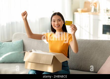 Online shopping concept. Excited asian woman receiving parcel, making YES gesture and showing credit card Stock Photo