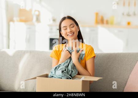 Excited young asian woman opening and unboxing package after delivery, hugging new shirt, sitting on couch and smiling Stock Photo