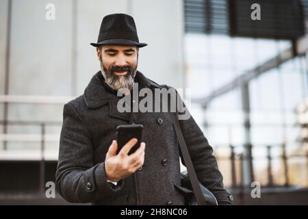 Portrait of happy businessman in black clothing standing in front of company building. Black bearded man is enjoying using smartphone. Stock Photo