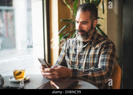 Adult man is sitting in cafe and messaging online. He is using smartphone. Stock Photo