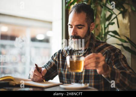 Adult man is sitting in cafe and making his daily schedule. He is drinking tea. Stock Photo