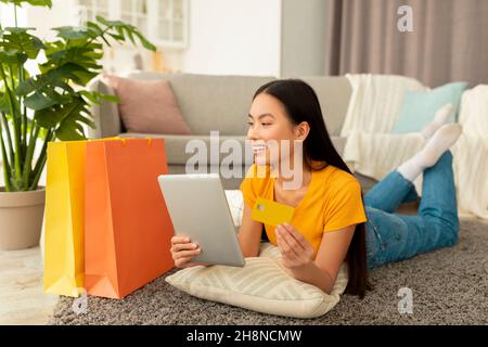 Shopaholism and e-commerce. Positive asian lady holding credit card, shopping online via tablet, lying on floor Stock Photo