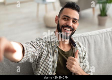 Happy young Arab guy taking selfie and showing thumb up gesture, smiling at mobile phone camera, sitting on sofa at home Stock Photo