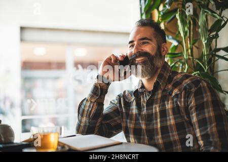 Adult man is sitting in cafe and making his daily schedule. He is talking on phone and using note pad. Stock Photo