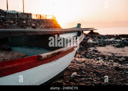 Old abandoned boat with rusted and chipped red and white paint, on a rocky beach at early morning sunrise golden hour Stock Photo