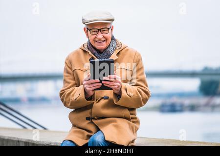 Outdoor portrait of cheerful senior man in winter coat who is using digital tablet. Stock Photo